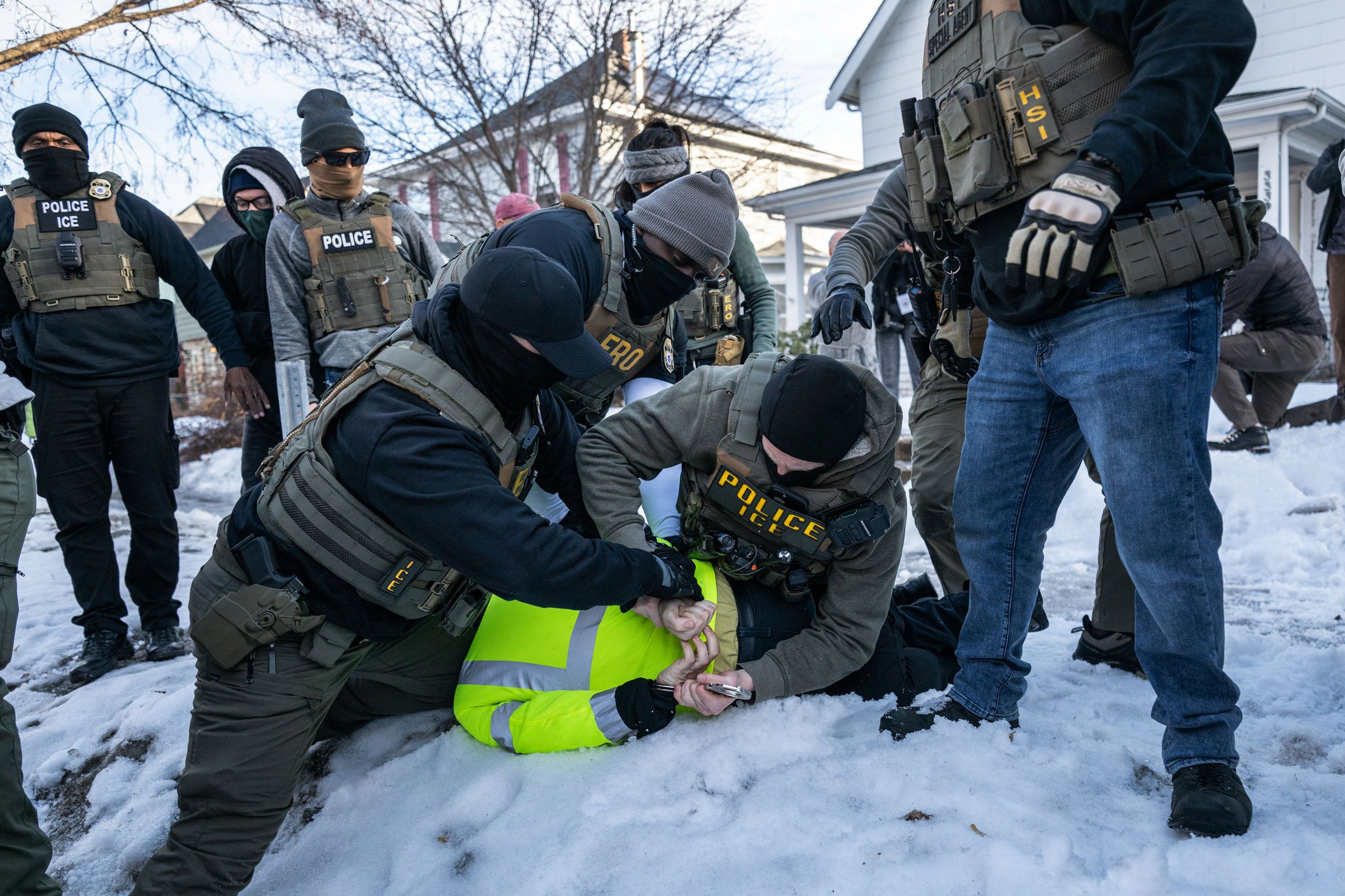 Several ICE agents restraining a person wearing yellow vest to the ground on snow.